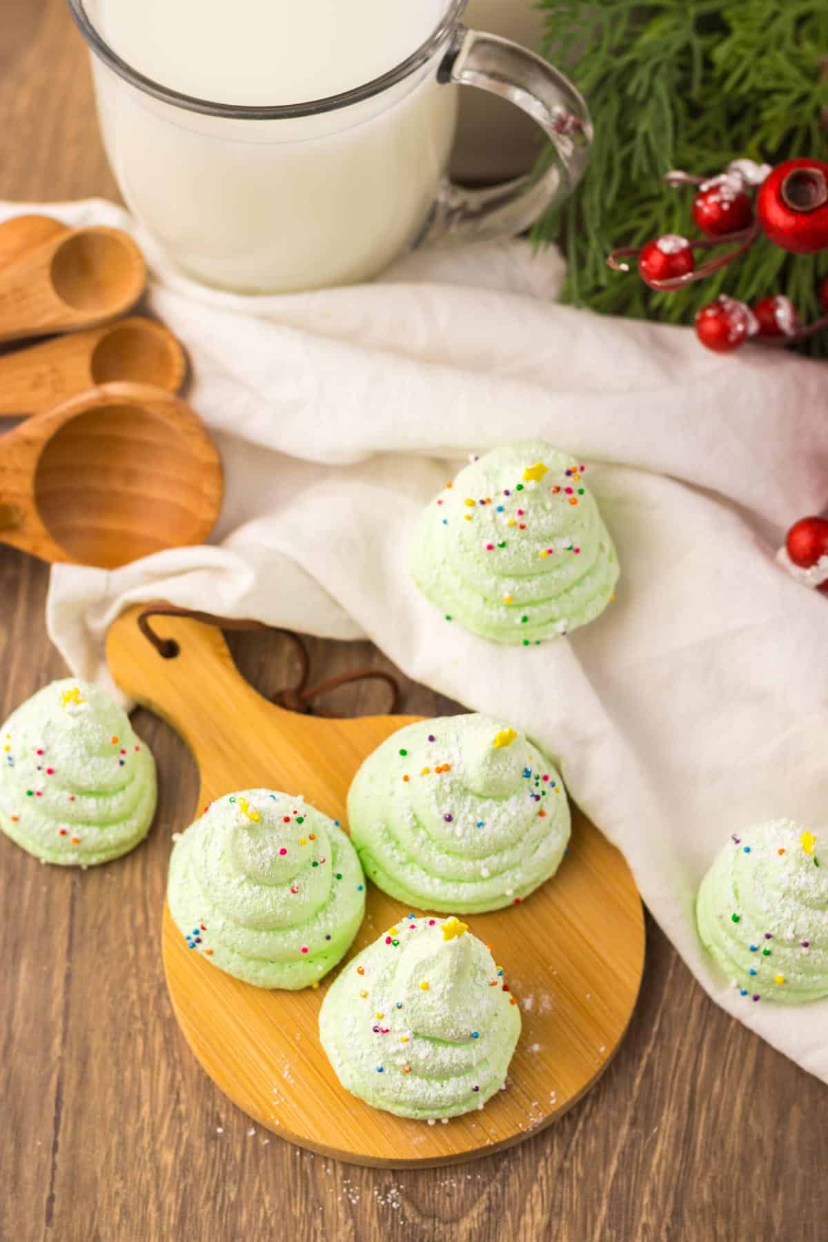 Green meringue cookies shaped like Christmas trees with sprinkles, on a wooden board next to a glass of milk.