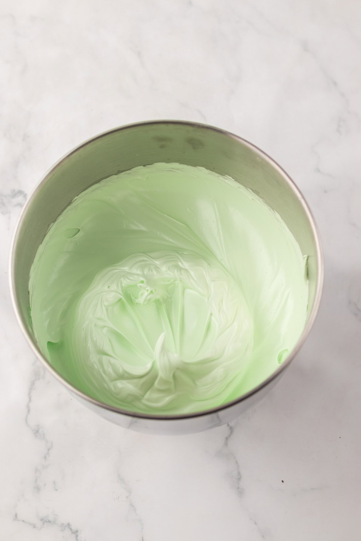 A metal bowl filled with light green, whipped cream or frosting on a marble surface.