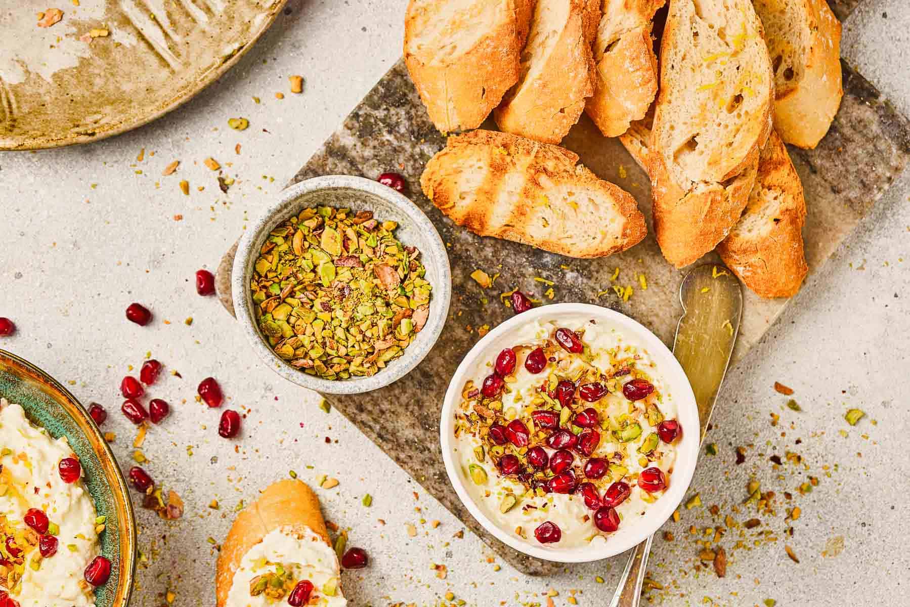 A bowl of Pomegranate Pistachio Whipped Feta dip topped with pistachios and pomegranate, surrounded by bread slices and a bowl of chopped pistachios.