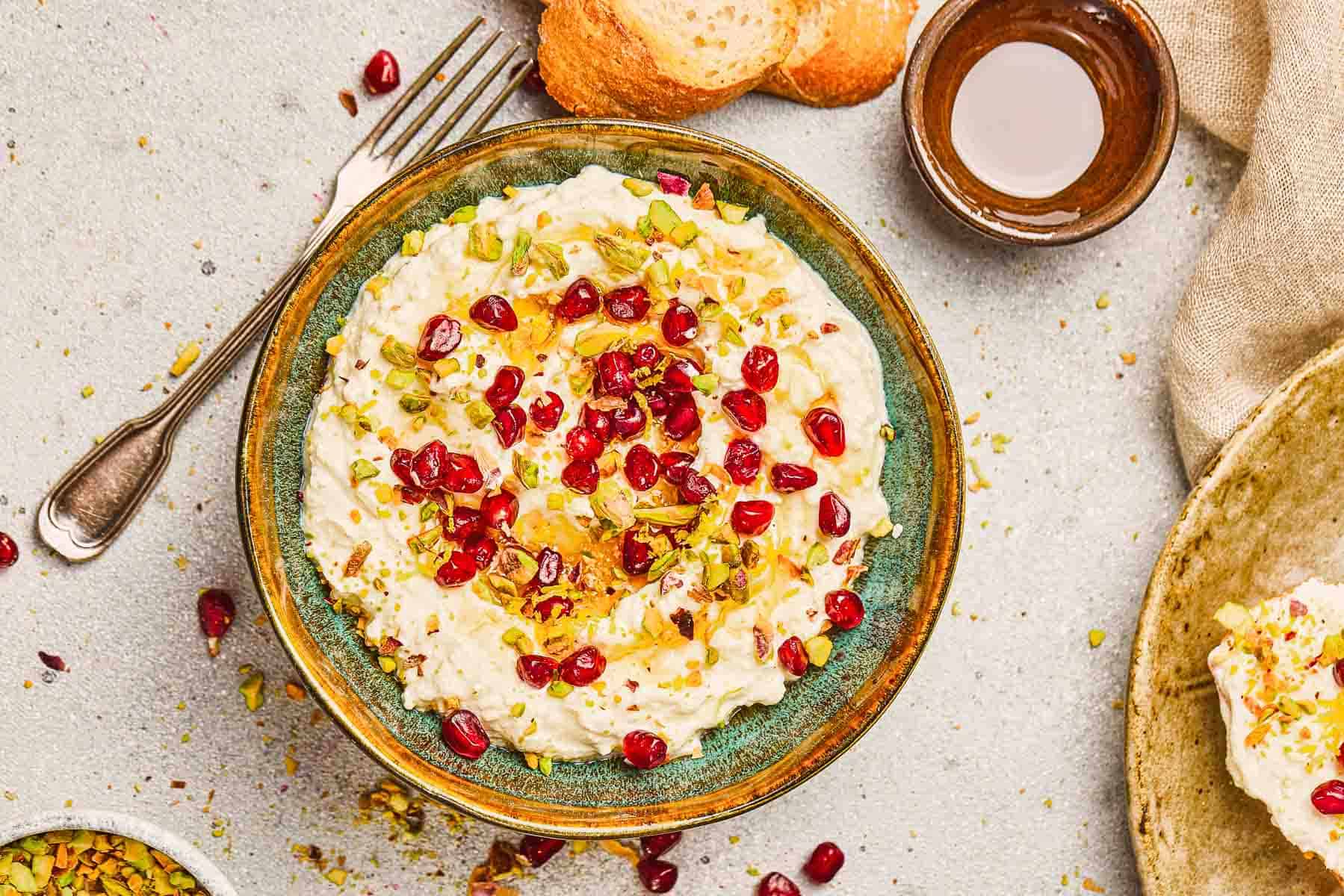A bowl of Pomegranate Pistachio Whipped Feta dip, topped with vibrant pomegranate seeds and chopped pistachios, served with bread slices nearby.