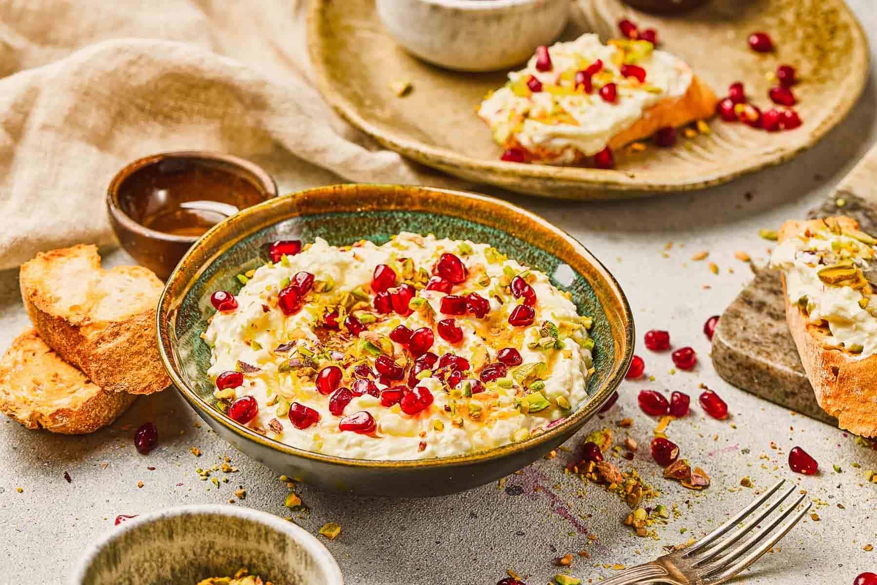 A bowl of creamy dip topped with pomegranate seeds and chopped nuts, with bread slices on the side.