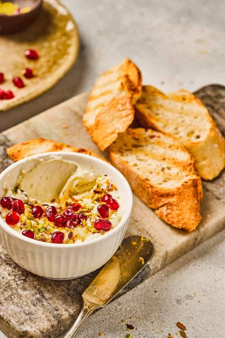 Sliced toasted bread with a bowl of Pomegranate Pistachio Whipped Feta dip, topped with pomegranate seeds and chopped nuts.