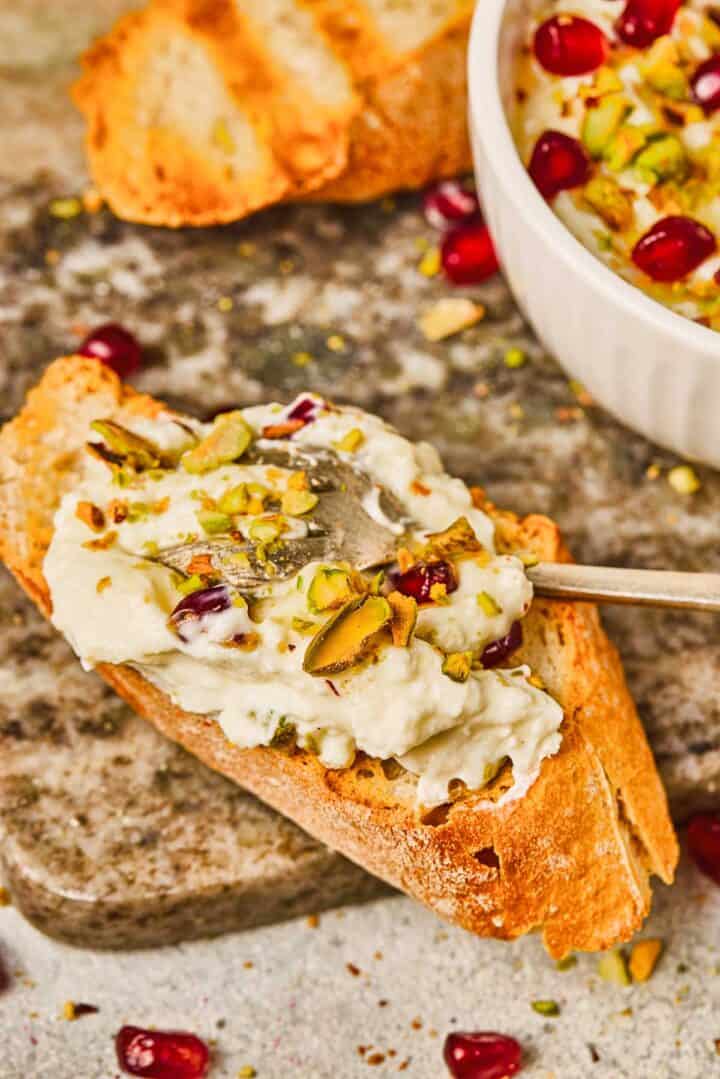 Toasted bread topped with pomegranate pistachio whipped feta and juicy pomegranate seeds, with a bowl of dip in the background.