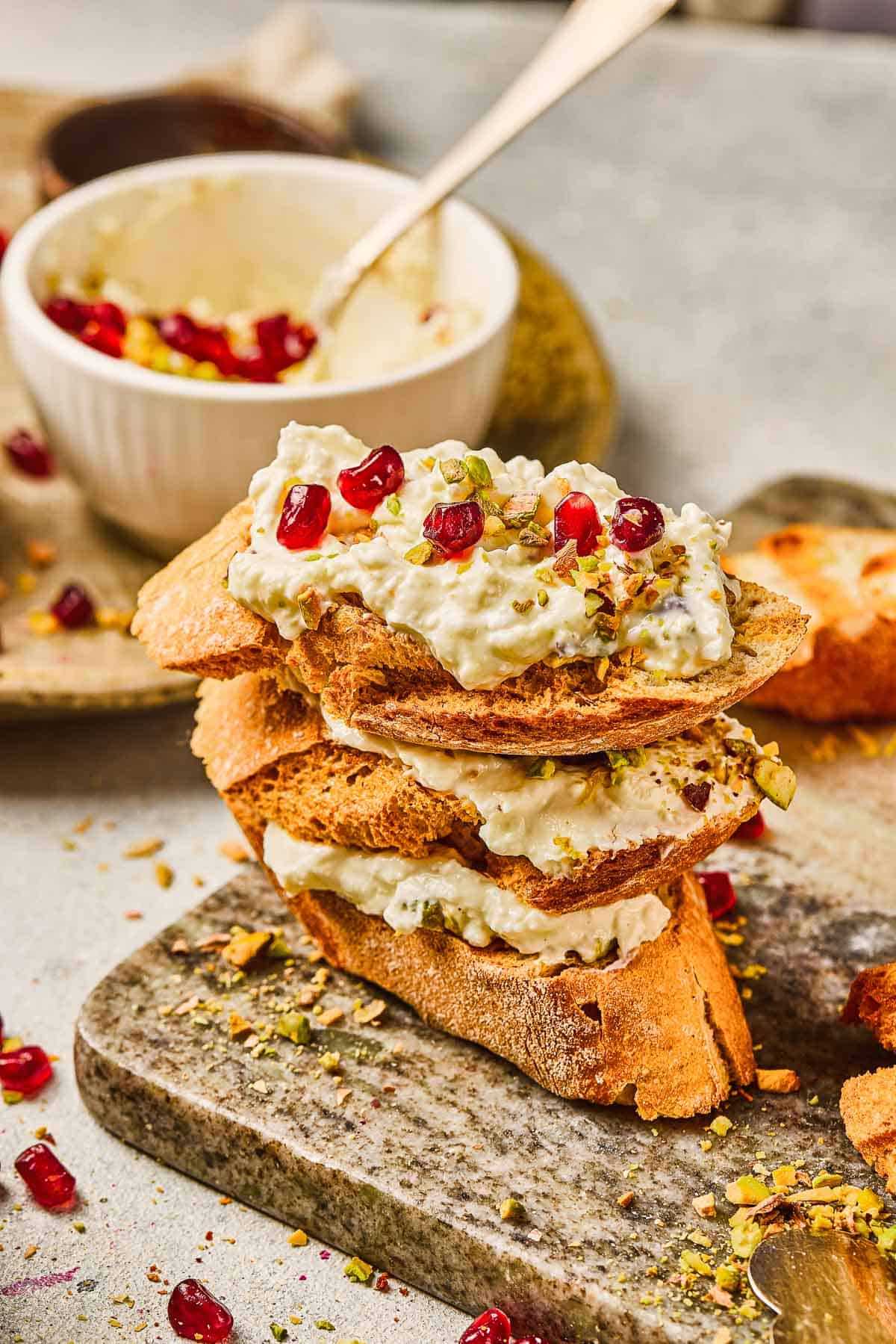 Three crostini topped with creamy cheese, pomegranate seeds, and chopped pistachios on a stone board.