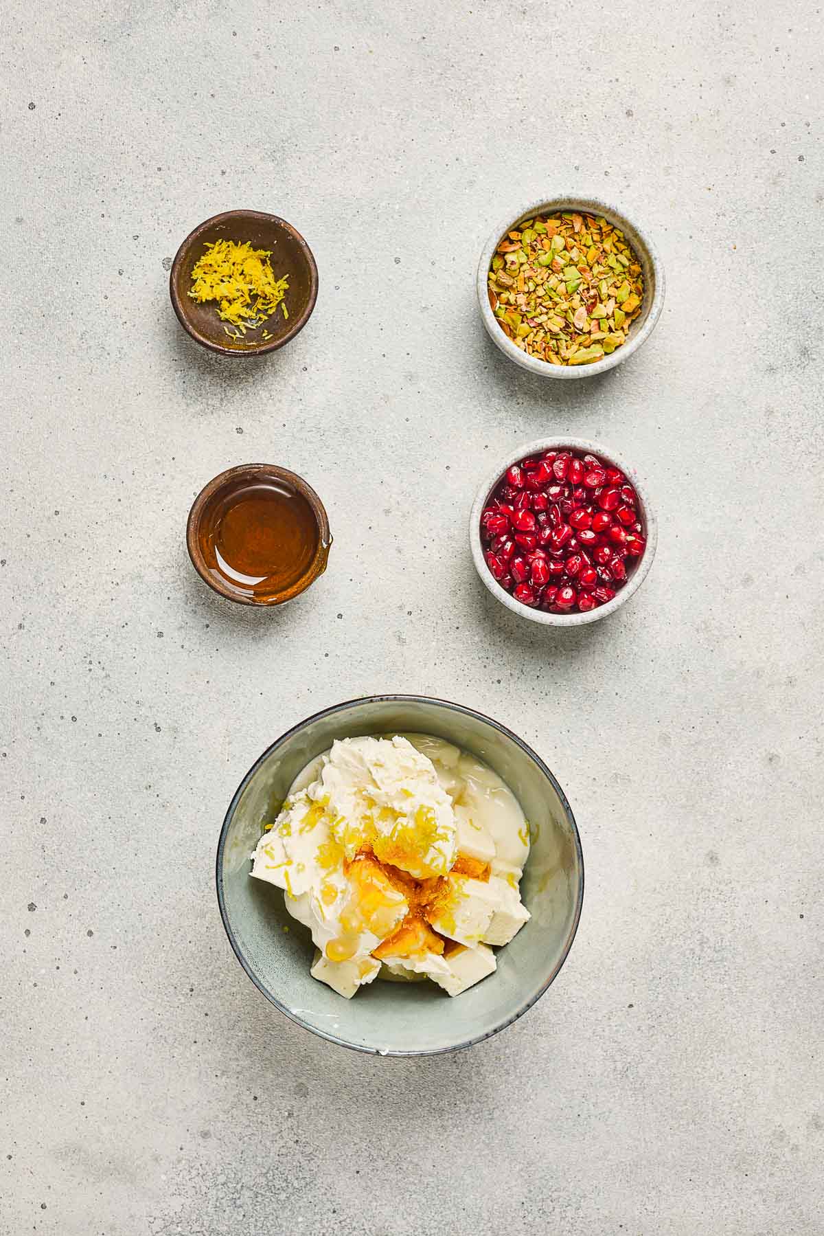 A bowl of pomegranate pistachio whipped feta with honey and cream cheese, surrounded by bowls of lemon zest, pistachios, pomegranate, and honey.