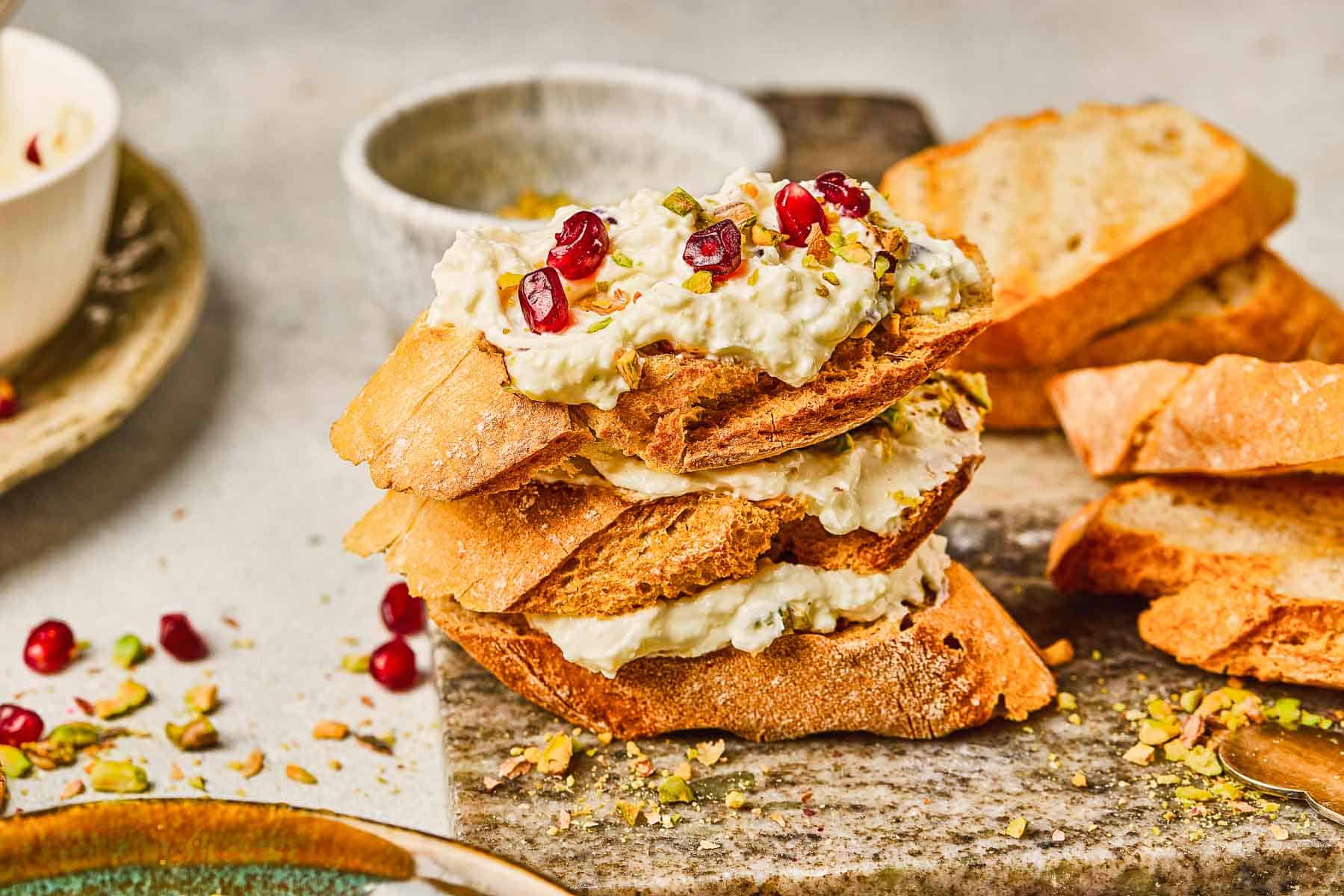 Toasted bread slices topped with creamy Pomegranate Pistachio Whipped Feta, chopped pistachios, and fresh pomegranate seeds on a board.