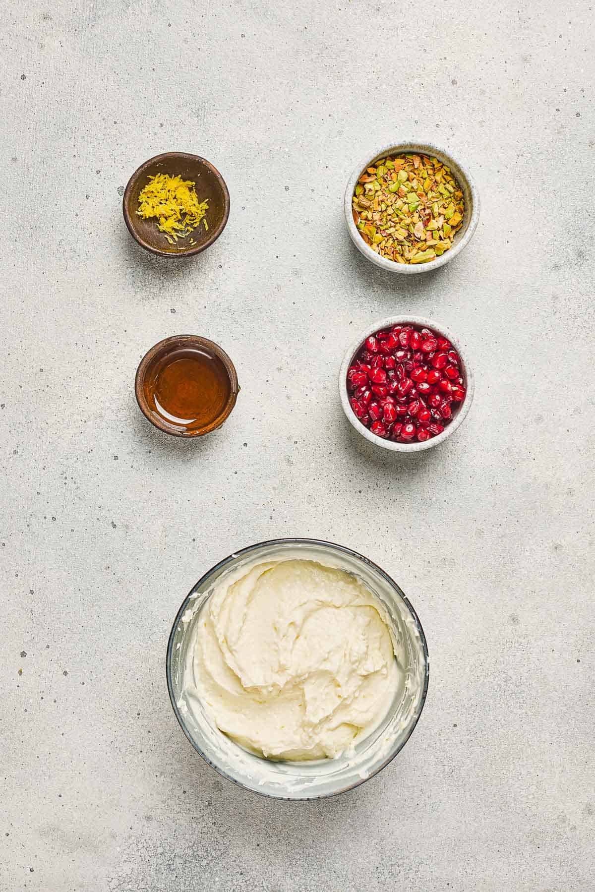 A bowl of Pomegranate Pistachio Whipped Feta with small bowls of lemon zest, honey, pistachios, and pomegranate seeds.