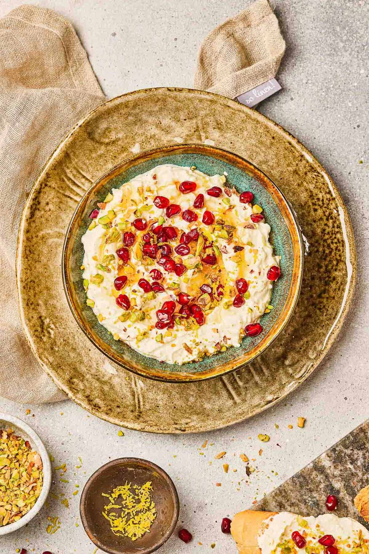 A bowl of rice pudding topped with pomegranate pistachio, whipped feta, and chopped pistachios on a rustic plate.