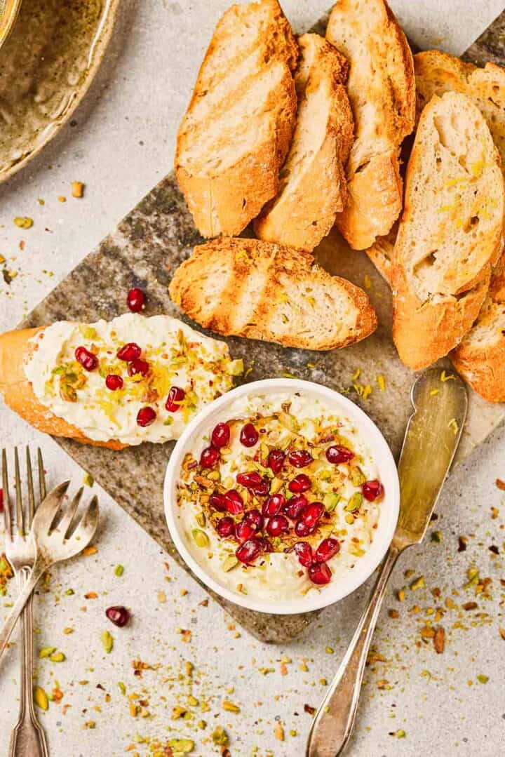 A bowl of Pomegranate Pistachio Whipped Feta topped with pomegranate seeds and nuts, served alongside grilled baguette slices on a cutting board.