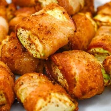 Golden brown bacon-wrapped zucchini pieces piled on a white plate, seasoned and roasted.