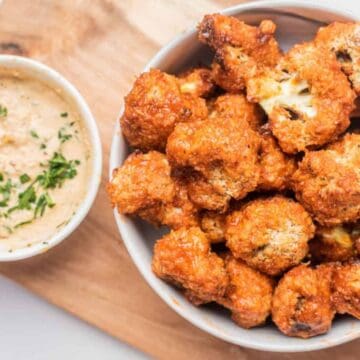 Bowl of crispy cauliflower bites with a side of creamy dipping sauce on a wooden board.