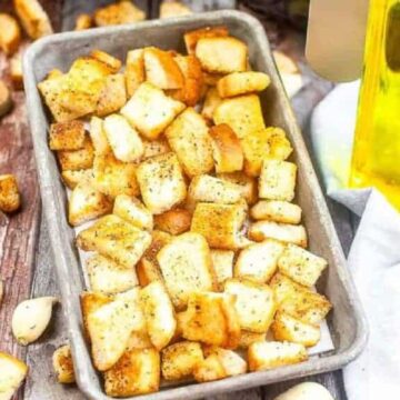 A tray of seasoned croutons next to a bottle of olive oil, bread slices, and garlic cloves.