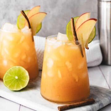 Two iced cocktails garnished with apple slices, lime, and cinnamon sticks on a marble board with a lime half.