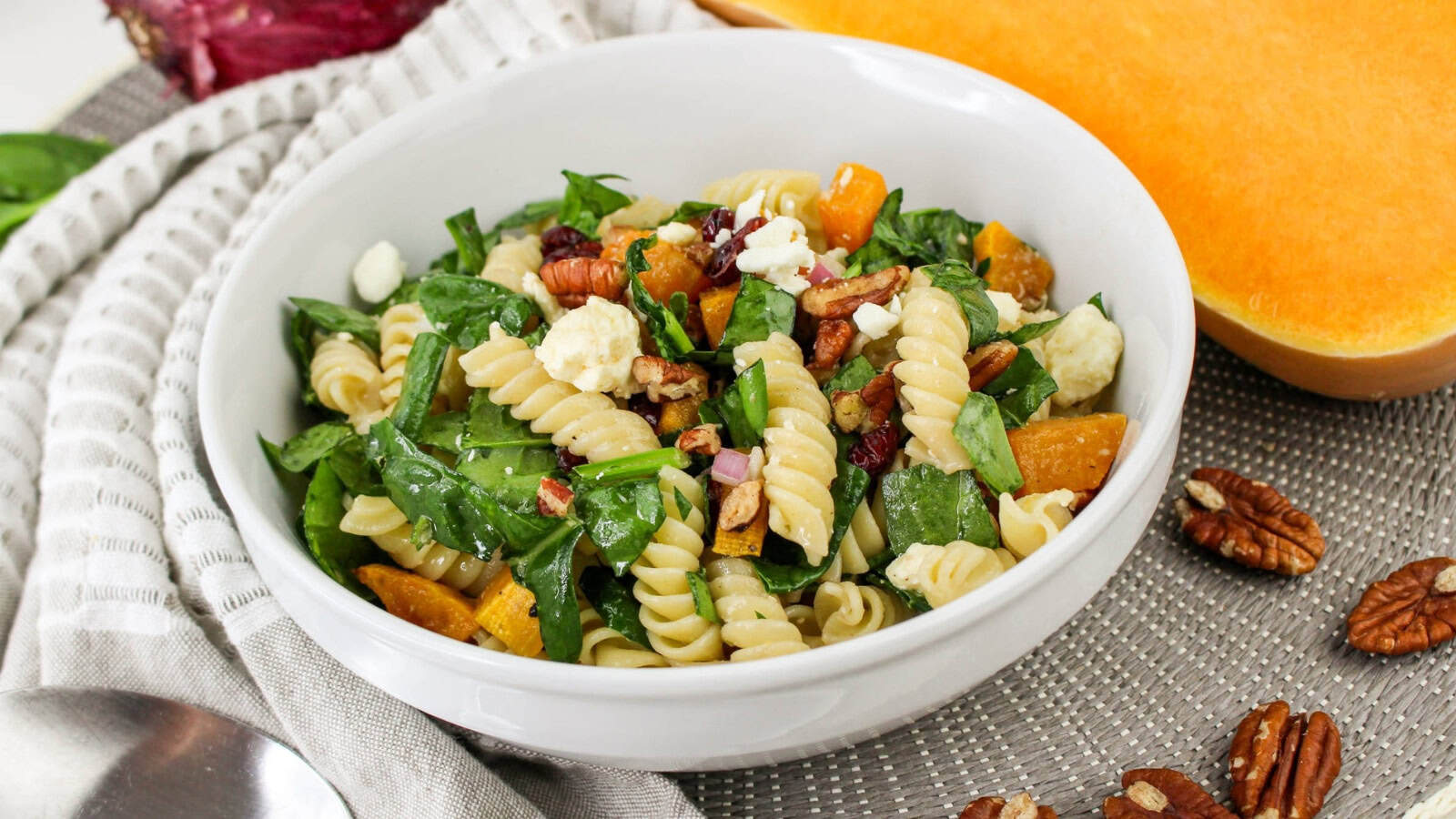 A bowl of fusilli pasta salad with spinach, roasted squash, pecans, and feta cheese, placed on a textured mat, with a spoon and sliced squash nearby.