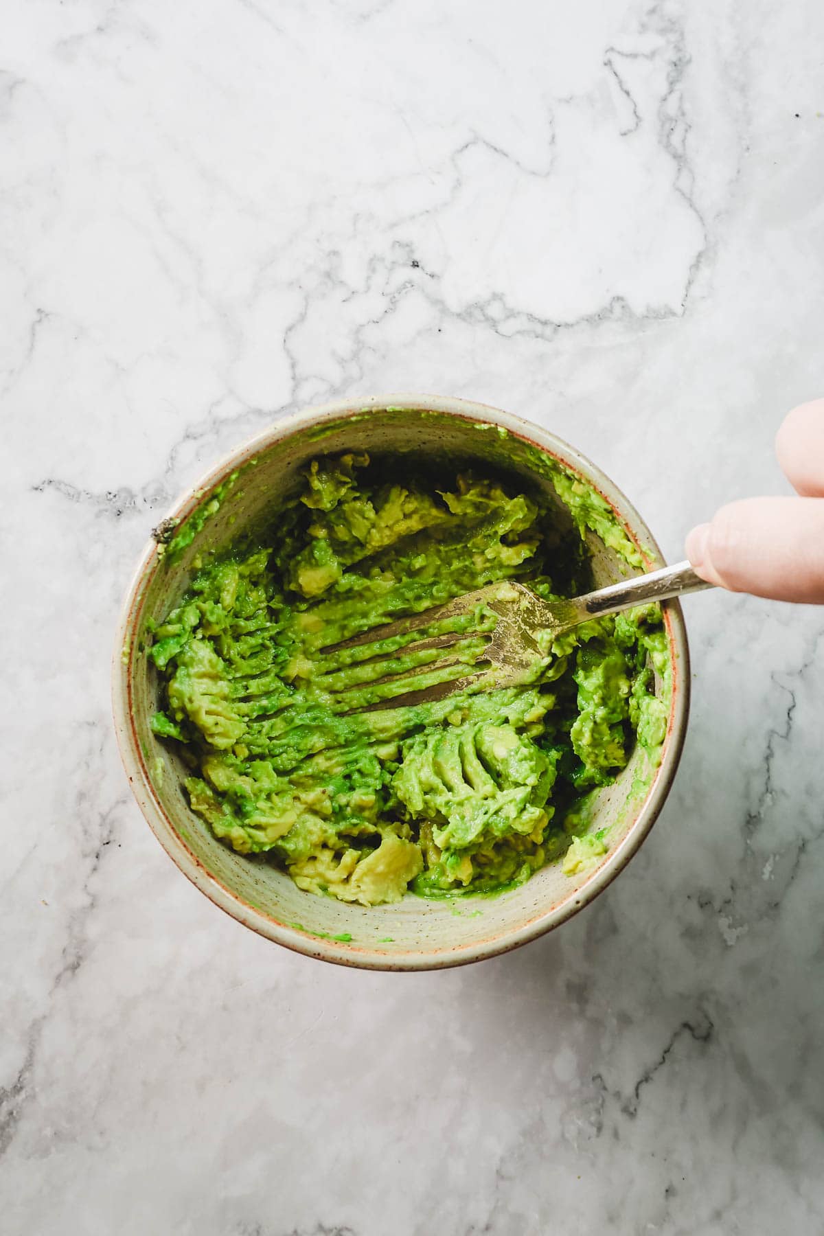 A hand mashes avocado with a fork in a bowl on a marble countertop, prepping for delicious Avocado Toast With Dukkah.