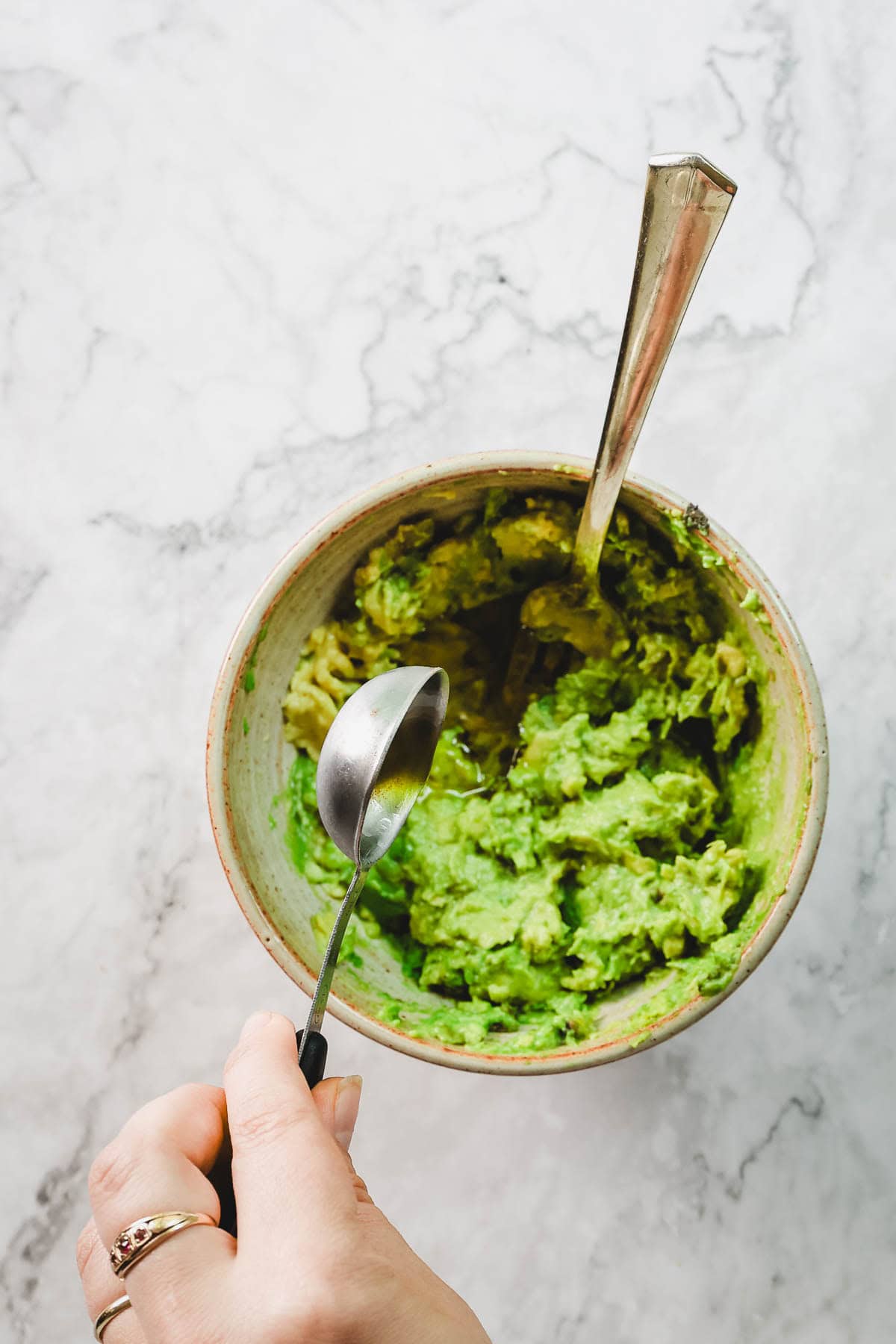 A hand holds a spoon over a bowl of mashed avocado with another spoon inside, on a marble surface-perfect for preparing Avocado Toast With Dukkah.