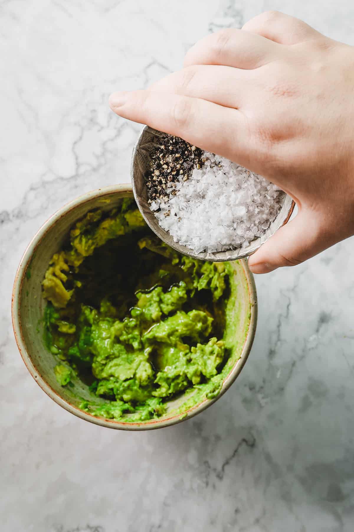 A hand adds salt and pepper to a bowl of mashed avocado on a marble countertop, preparing the base for Avocado Toast With Dukkah.
