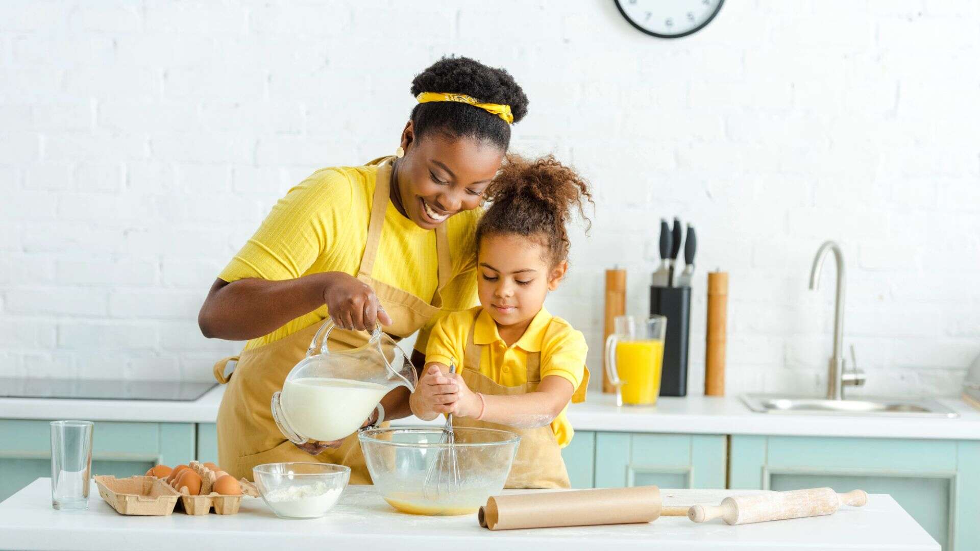 Smiling woman and girl in yellow aprons use kitchen tools for holiday baking as they mix ingredients and pour milk in a bright kitchen.