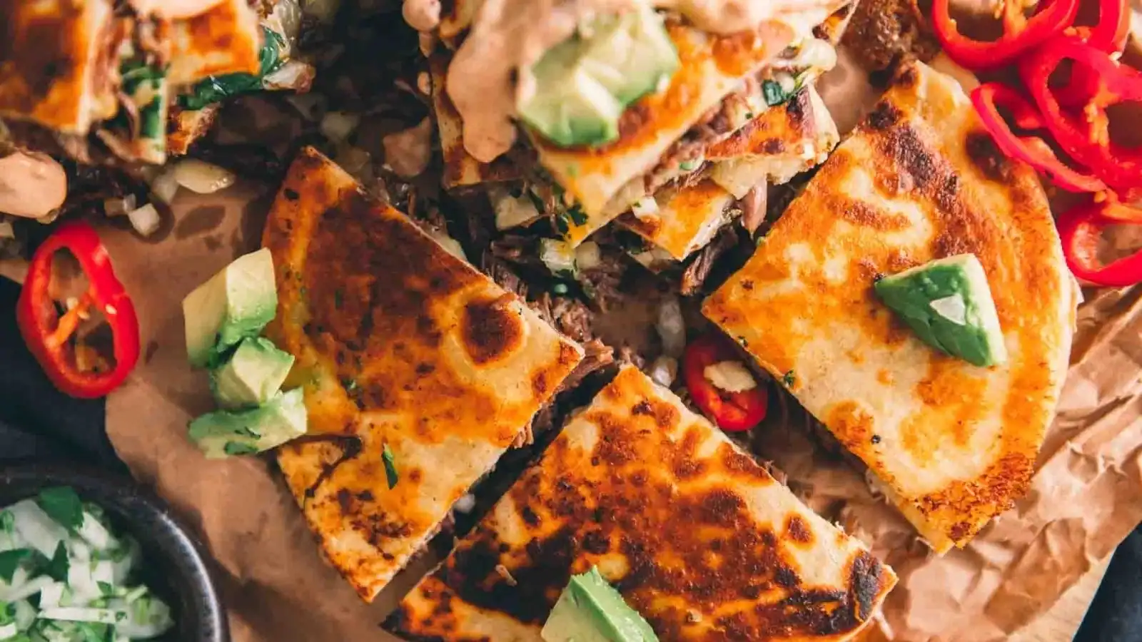 Golden, crispy quesadillas topped with avocado and sliced red chili peppers on parchment paper.