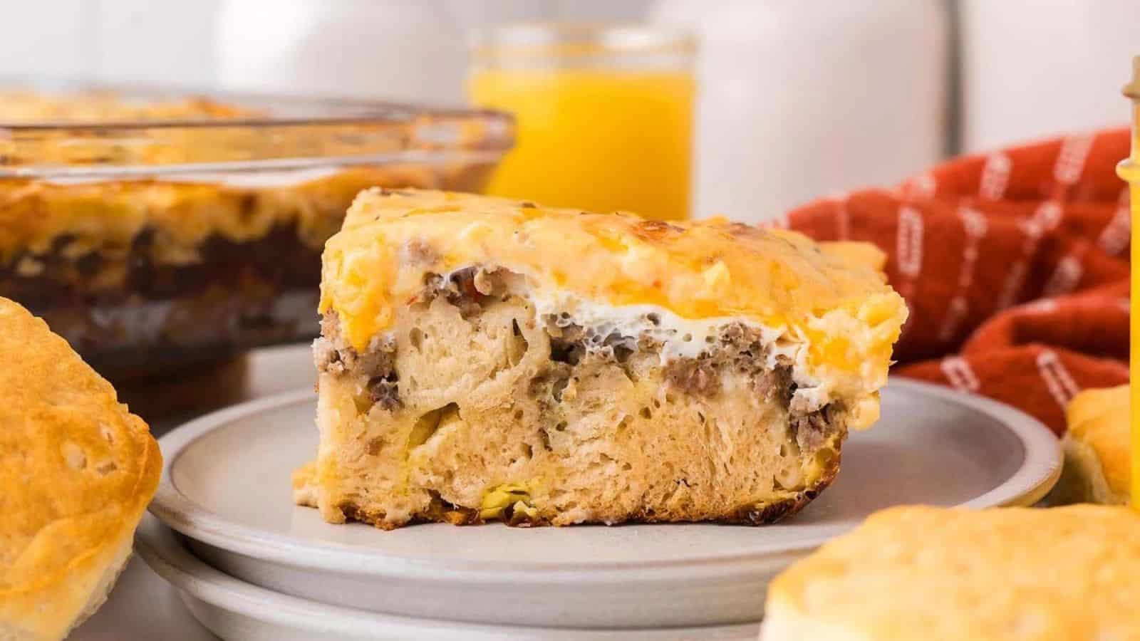 A slice of breakfast casserole with sausage, egg, and cheese on a plate, with biscuits and orange juice nearby.