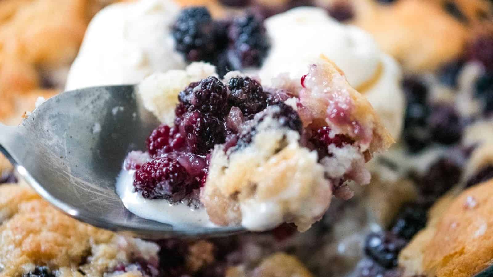 A cast iron skillet with a baked berry cobbler topped with a scoop of vanilla ice cream and garnished with fresh blackberries.