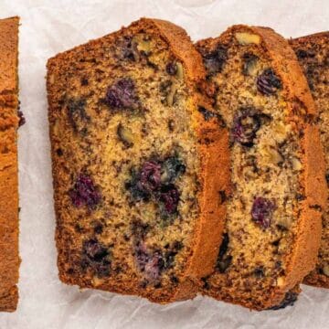 Three slices of blueberry nut bread next to a loaf on white parchment paper, showing berries and nuts inside.