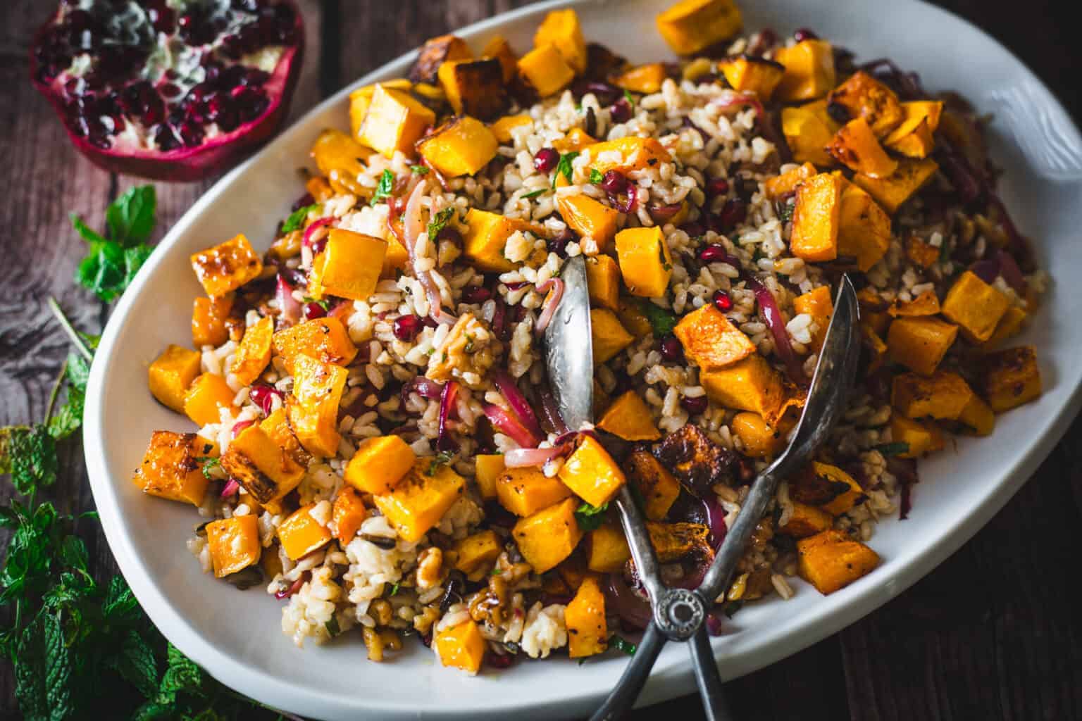 Oval dish with roasted butternut squash, rice, red onions, herbs, and pomegranate seeds; serving tongs on side.
