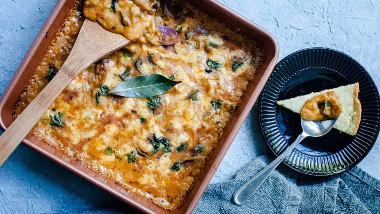 Baked casserole with melted cheese and herbs in a square dish, a slice served on a black plate nearby.