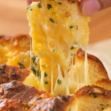 A hand pulls cheesy, herb bread apart, with melted cheese stretching between the pieces.