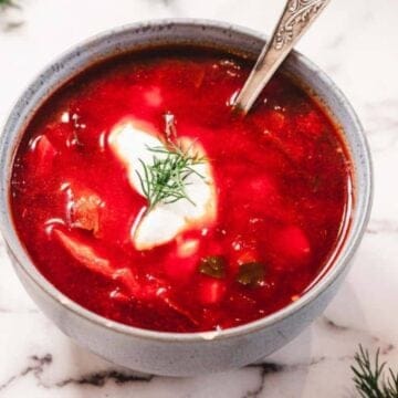 A bowl of red soup with a dollop of sour cream and dill, on a white marble surface with herb sprigs nearby.