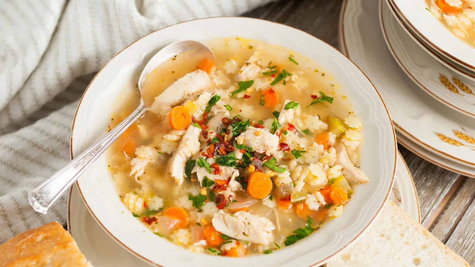 A bowl of chicken soup with pasta, carrots, and herbs, served with a slice of bread on the side.