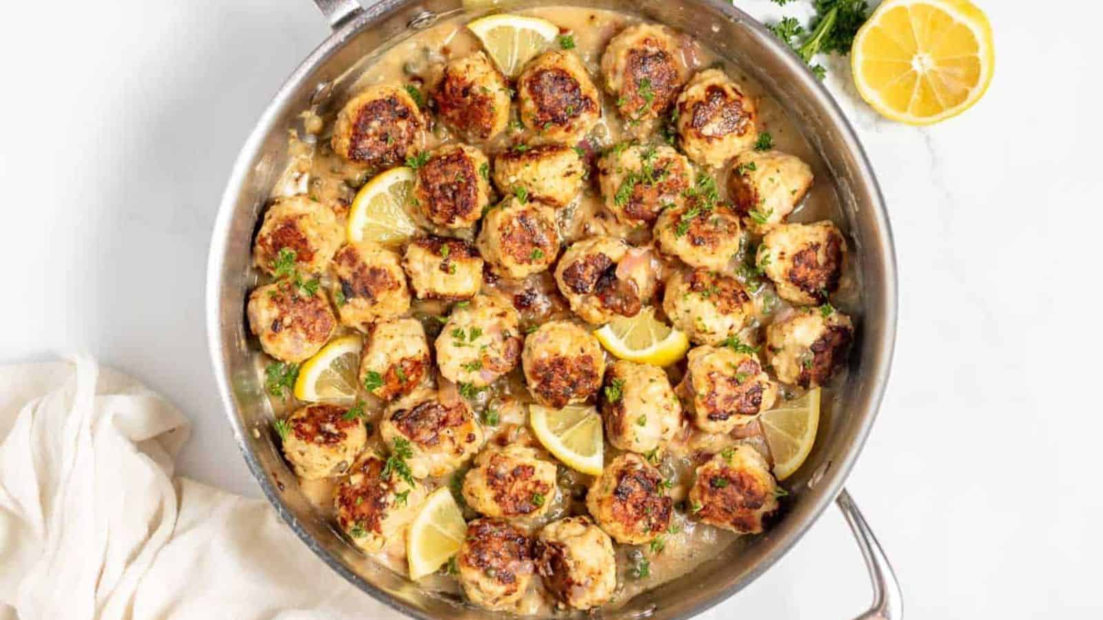 A skillet with cooked browned meatballs in a lemon herb sauce, garnished with chopped parsley and lemon wedges. A white cloth and a cut lemon are placed beside the skillet on a white surface.