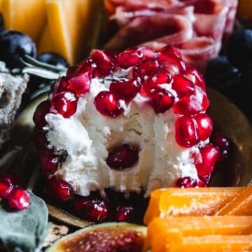 A round cheese ball covered with pomegranate seeds on a platter with meats, fruits, and cheese.
