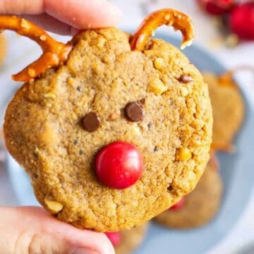 A hand holds a reindeer-shaped cookie with pretzel antlers, chocolate eyes, and a red candy nose.