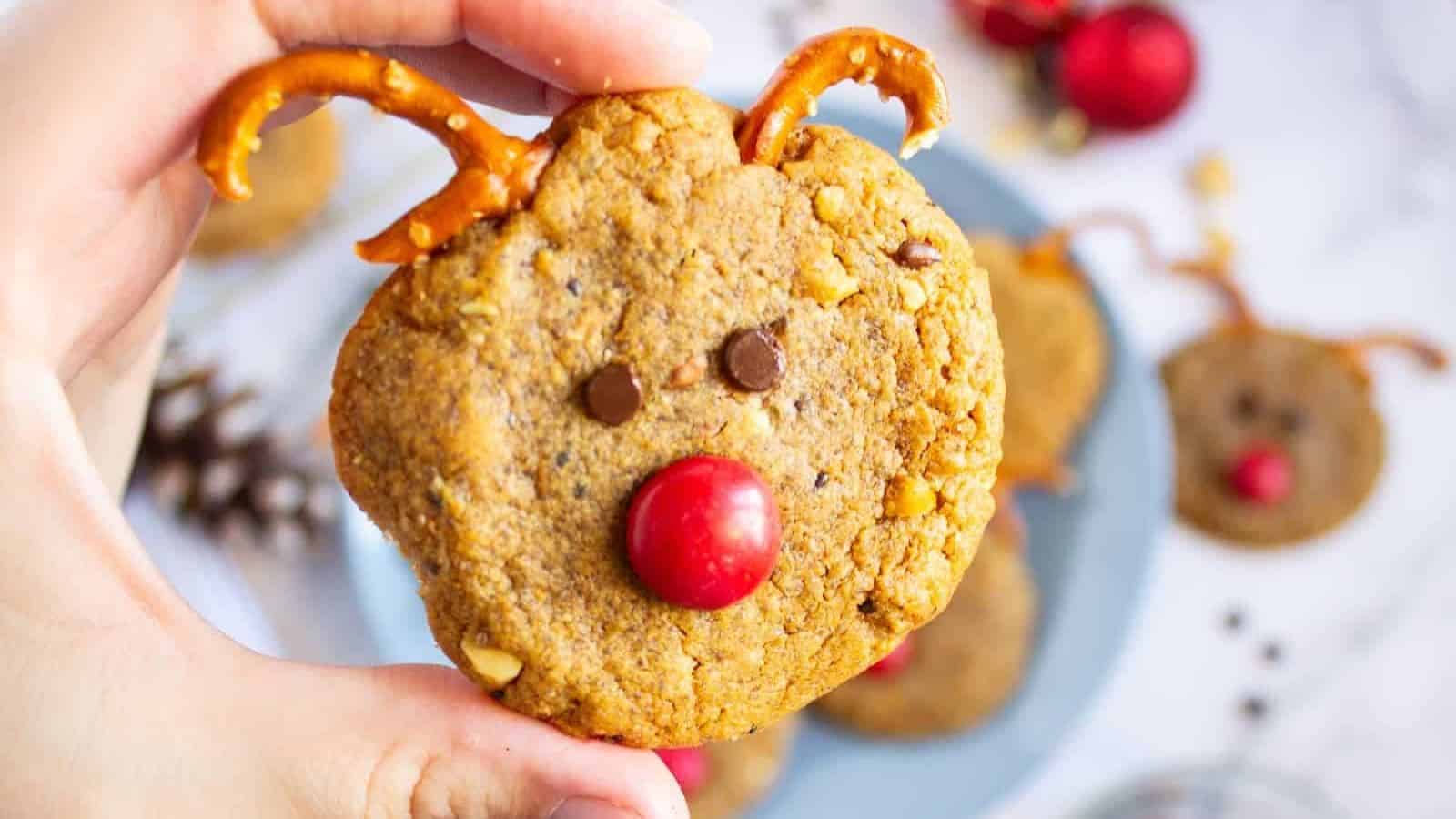 A plate of Christmas reindeer cookies with pretzel antlers, chocolate chip eyes, and red M&M noses, surrounded by holiday decorations and baking ingredients.
