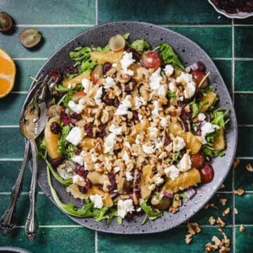 Arugula salad with orange slices, grapes, walnuts, cranberries, and goat cheese on a gray plate.