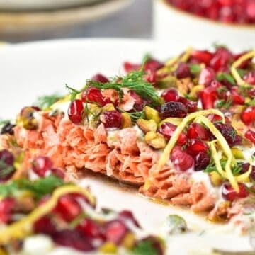 Close-up of baked salmon topped with pomegranate seeds, lemon zest, herbs, and a creamy sauce on a white plate.