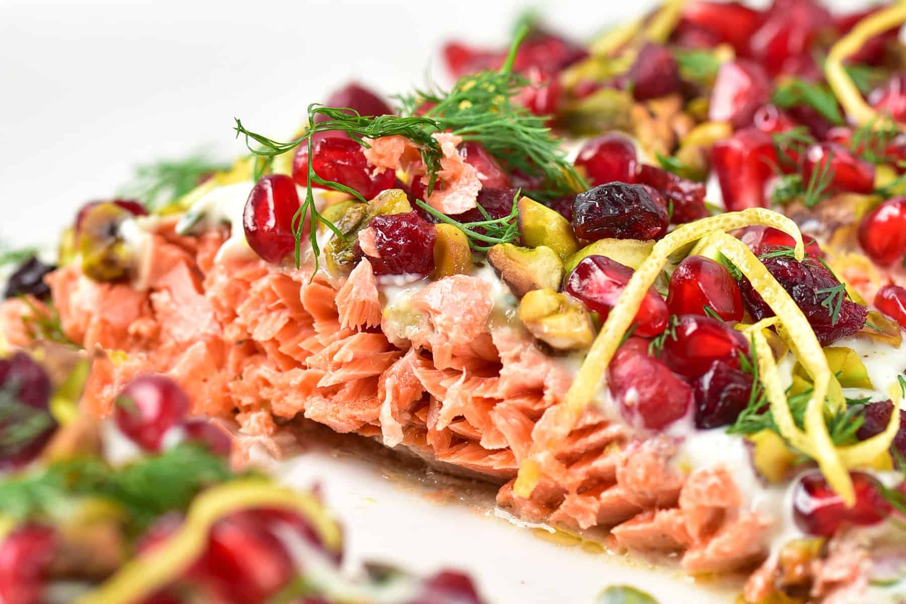 Close-up of baked salmon topped with pomegranate seeds, pistachios, lemon zest, fresh dill, and creamy sauce.