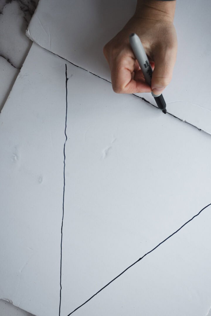 A hand draws black lines on a white foam board with a black marker, sketching out the outline for a festive Christmas Tree Charcuterie Board design.