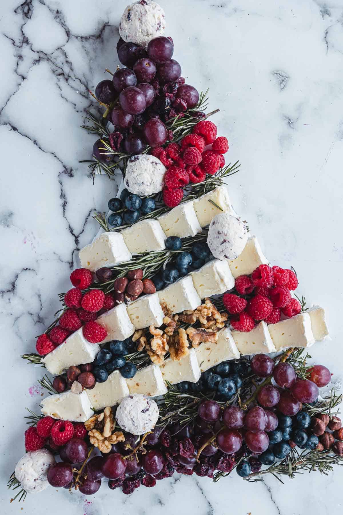Christmas Tree Charcuterie Board featuring grapes, raspberries, blueberries, cheese, nuts, and rosemary arranged on a marble base.