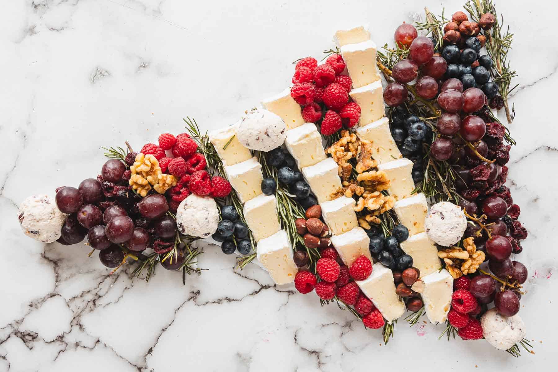 Cheese, grapes, berries, nuts, and herbs are artfully arranged in a Christmas Tree Charcuterie Board shape on a marble surface.