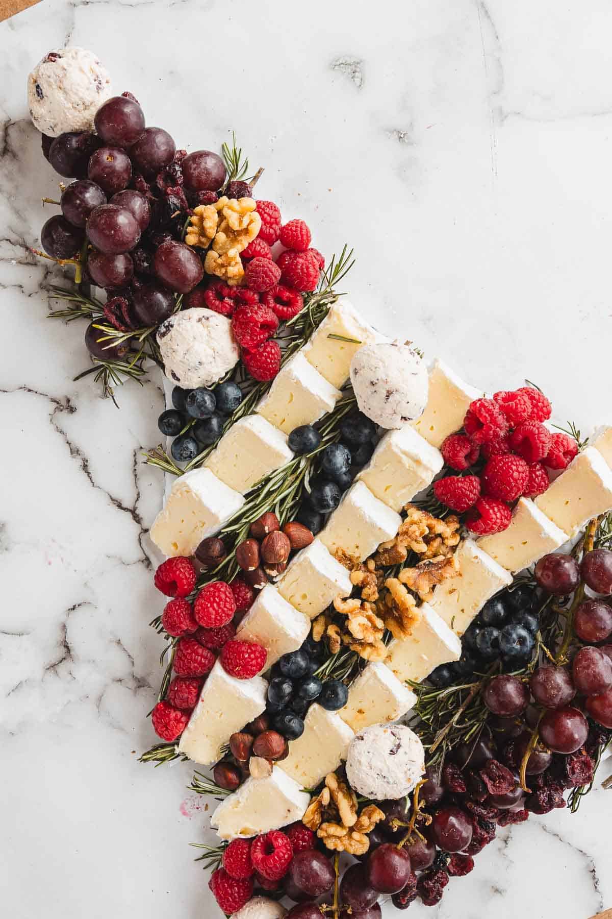 Diagonal cheese board with brie, grapes, berries, nuts, and herbs on a white marble surface&mdash;perfect inspiration for your next Christmas Tree Charcuterie Board.