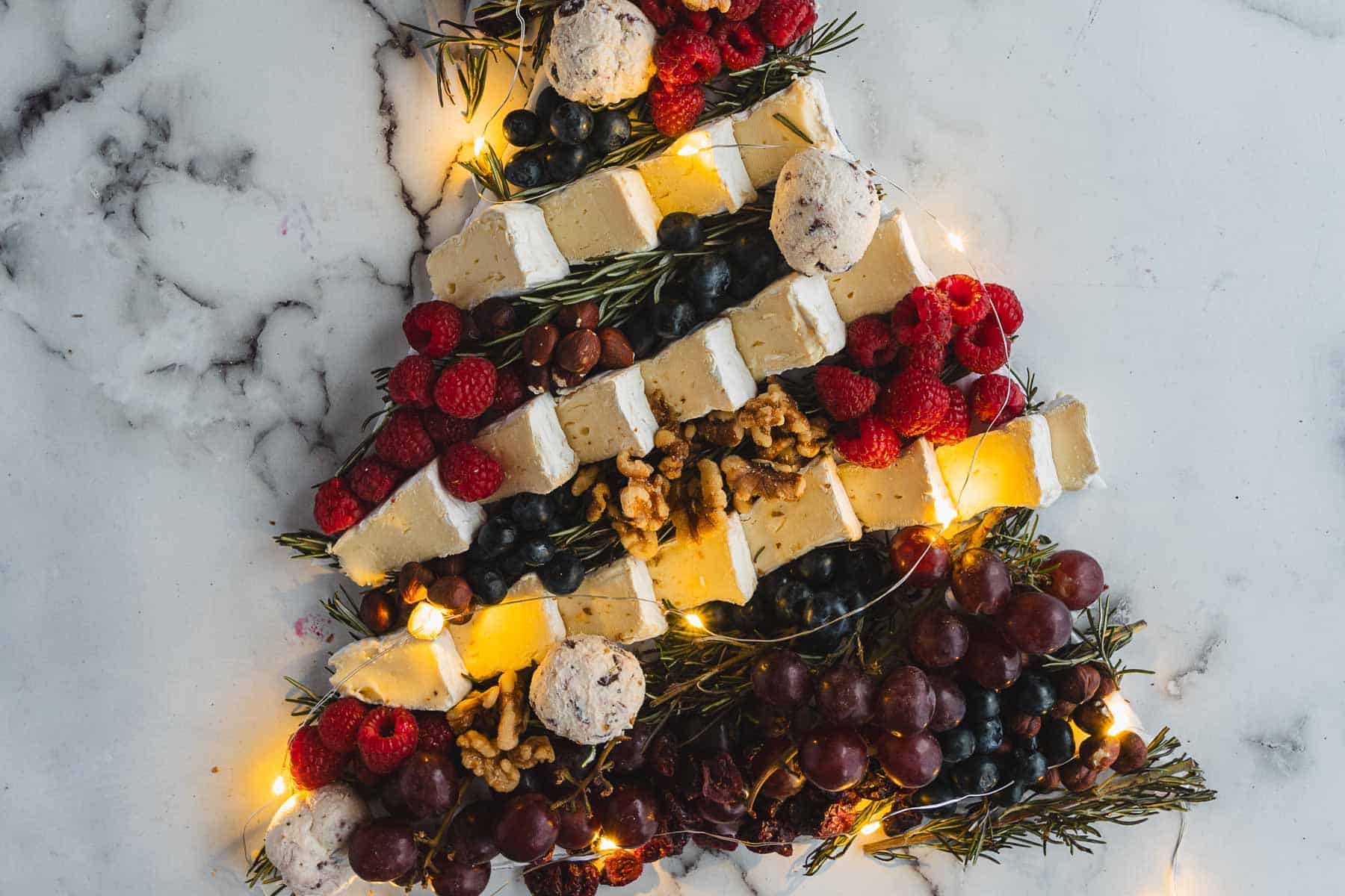 Christmas Tree Charcuterie Board arranged with cheese, berries, grapes, walnuts, rosemary, and twinkling string lights for a festive touch.