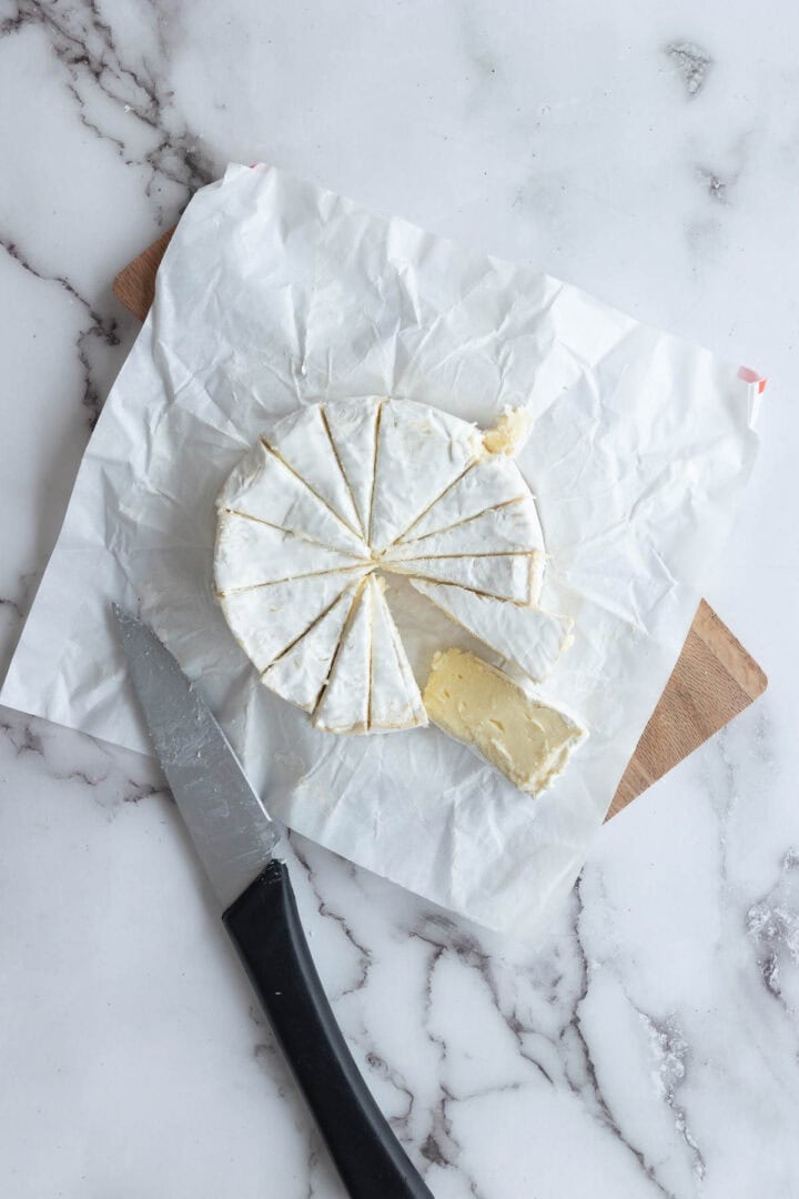 A wheel of brie cheese cut into wedges sits on parchment paper with a knife beside it, ready to be added to your festive Christmas Tree Charcuterie Board on a marble surface.