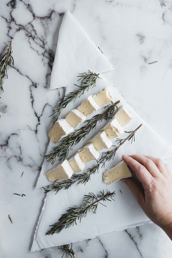 Hand placing cheese slices on parchment shaped like a Christmas tree, creating a festive Christmas Tree Charcuterie Board garnished with rosemary sprigs.