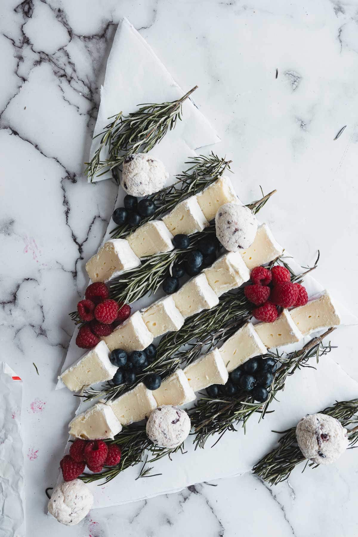 Sliced brie, berries, and rosemary are artfully arranged as a Christmas Tree Charcuterie Board on marble, with cheese balls as festive ornaments.