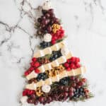 Christmas Tree Charcuterie Board arranged with cheese, berries, grapes, nuts, and herbs on a marble background.