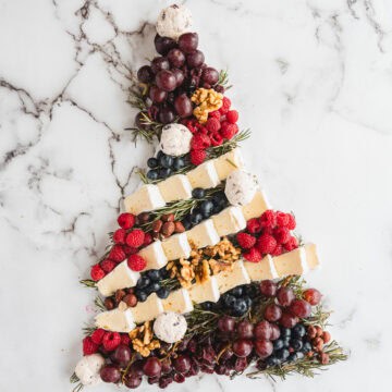 Christmas Tree Charcuterie Board arranged with cheese, berries, grapes, nuts, and herbs on a marble background.