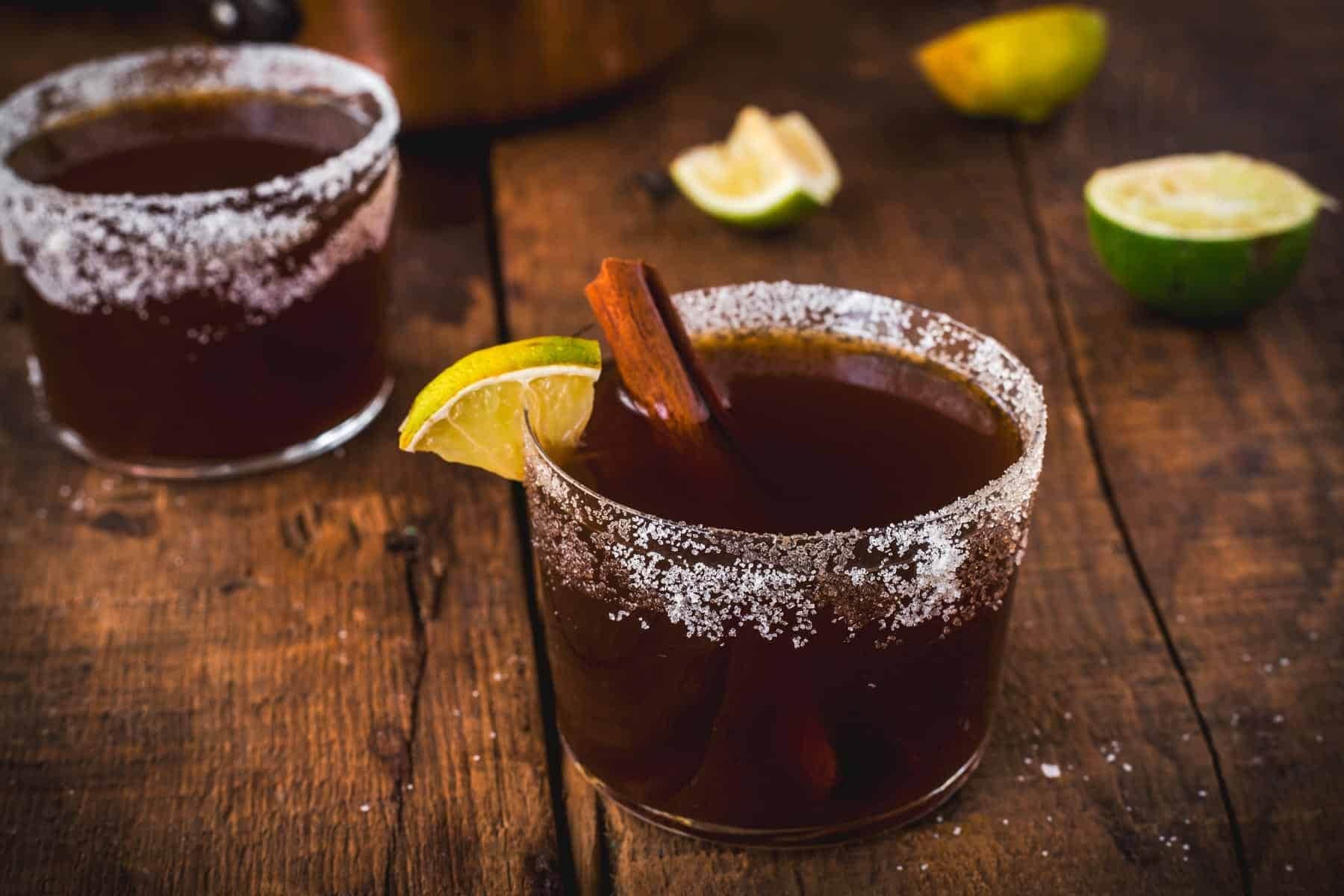 A glass of dark beverage with a salted rim, lime wedge, and cinnamon stick on a rustic wooden table.