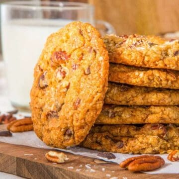 A stack of oatmeal cookies with pecans sits on a wooden board next to a glass of milk.